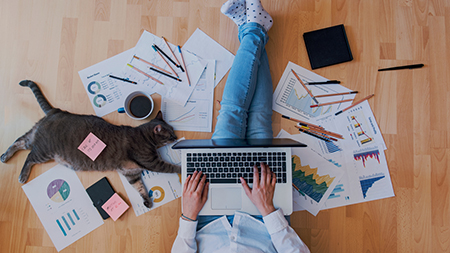 En person jobber på gulvet omgitt av papir, pc, kaffekopp og en katt