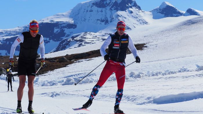 En trener og en skiløper går på ski i solskin med fjell bak seg. 