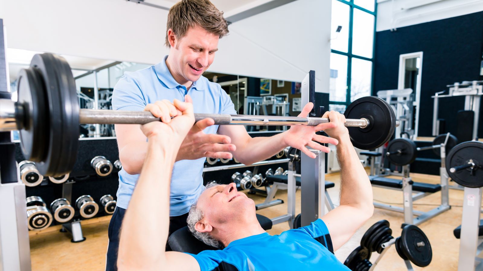 Personlig trener/fysiolog som hjelper en eldre mann med å løfte vekter/benkpress på et treningssenter.